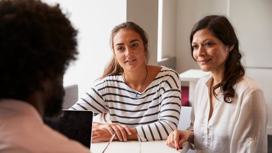 two women sit at a desk in an office facing a man and his laptop